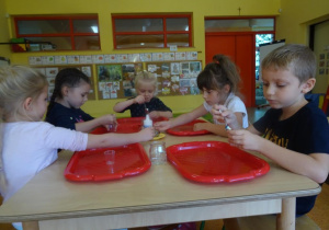 Dzieci siedzą przy stoliku i za pomocą łyżeczek przesypują drożdże z talerzyka na środku stolika do swoich małych plastikowych pojemników.